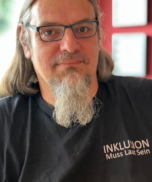 Ron Paustian is responsible for inclusion and accessibility. He has long curly hair, wears glasses and has a long chin beard. The name Inklusion Muss Laut Sein (Inclusion Must Be Loud) can be read on his T-shirt.